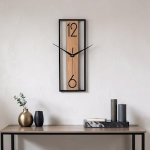 May include: A rectangular wall clock featuring a black metal frame surrounding a vertical light brown wooden panel. The clock face displays the numbers 12 and 6 in a black sans-serif font, with two thin black metal hands centered on the panel.