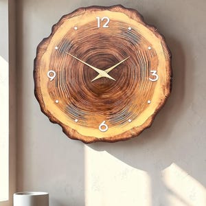May include: A round wall clock shaped like a natural wood slice with visible tree rings. The clock face features a light brown center, dark brown edges, white numerals at 12, 3, 6, and 9, small white dot markers, and two thin gold-colored hands.