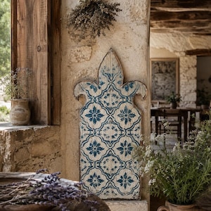 May include: A tall, vertical wooden decorative panel featuring a pointed, fleur-de-lis inspired top. The surface is painted with a distressed white finish and a repeating blue floral tile pattern, showing visible wood grain and weathered edges.
