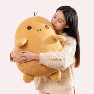 Op de afbeelding: Een grote, goudbruine knuffel in de vorm van een aardappel met een zacht, getextureerd oppervlak. Het heeft twee kleine, donkerbruin geborduurde ogen, een eenvoudig gestikte mond, twee korte armpjes, twee beentjes en een klein spruitje bovenop.