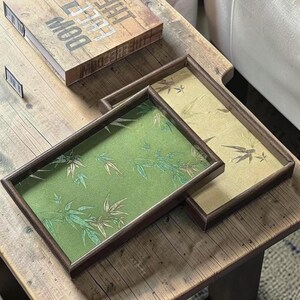 Peut inclure: Deux plateaux rectangulaires en bois avec des cadres marron foncé. Le plateau supérieur présente une incrustation en tissu vert avec un motif de feuilles de bambou, tandis que le plateau inférieur a une incrustation beige avec un motif similaire.