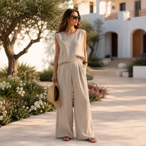 Op de afbeelding: Een beige set bestaande uit een mouwloze top en een bijpassende broek met wijde pijpen van gestructureerde stof. De outfit wordt gecombineerd met bruine platte sandalen en een strooien hoed in de hand. Het geheel heeft een eenvoudige snit.