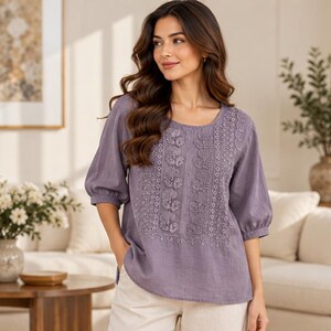 Pode incluir: Uma blusa em tom roxo poeirento com mangas bufantes de três quartos e decote redondo. O painel frontal é decorado com filas verticais de bordados florais tonais e detalhes de costura festonada sobre uma base de tecido liso.