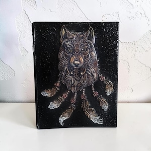 Könnte beinhalten: Ein rechteckiger schwarzer Bucheinband mit einem erhabenen, metallisch wirkenden Relief eines Wolfskopfes mit komplexen geometrischen Mustern. Unter dem Wolf sind mehrere herabhängende Federn und kleine kreisförmige Akzente in Bronze- und Silbertönen dargestellt.