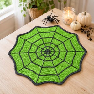 May include: A round, crocheted doily featuring a vibrant lime green spider web pattern outlined in dark charcoal gray yarn. The circular textile has a scalloped edge and a geometric web design radiating from the center to the outer border.