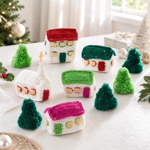 Op de afbeelding: Een verzameling gehaakte kerstversieringen, waaronder verschillende kleine huisjes met rode, groene en roze daken, een witte kerk met een toren en diverse kegelvormige kerstbomen van licht- en donkergroen garen.