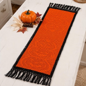 May include: A rectangular orange crochet table runner featuring a repeating pumpkin pattern, bordered by a black crochet edge and finished with black fringe at both ends. The runner is displayed alongside a small orange pumpkin and dried leaves.
