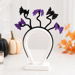 May include: A black headband featuring five thin, flexible wire stems topped with crocheted bat shapes in alternating black and purple colors. The bats are arranged in a fan-like pattern, with each stem extending upward from the main headband base.