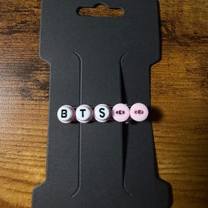 Pode incluir: Um gancho de cabelo com três contas circulares brancas com as letras pretas B, T e S impressas, seguidas por duas contas circulares rosa claro com pequenos orifícios no centro, montado num cartão de apresentação retangular preto.