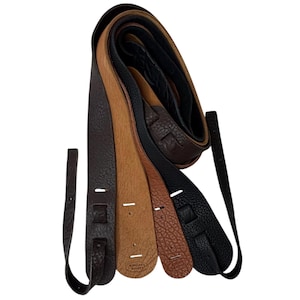 May include: A collection of four guitar straps in varying shades of brown and black, featuring a textured surface. Each strap includes reinforced ends with slots for attachment, and the two center straps display the embossed text Dakota Leathers.
