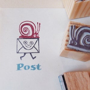 Puede incluir: Un conjunto de tres sellos de madera con diseños de caracoles. Los sellos están montados en bloques de madera. Un sello muestra un caracol llevando una carta con la palabra "Post" impresa debajo.