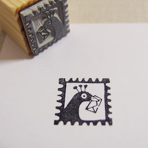May include: A black and white rubber stamp featuring a peacock design. The peacock is holding a letter in its beak. The stamp is in a square frame with a scalloped edge.