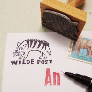 Pode incluir: Um carimbo de madeira com um desenho de hiena de desenho animado, o texto "WILDE POST" e as letras vermelhas "An" são estampados em papel branco. Um selo postal de elefante e uma caneta preta também são visíveis.