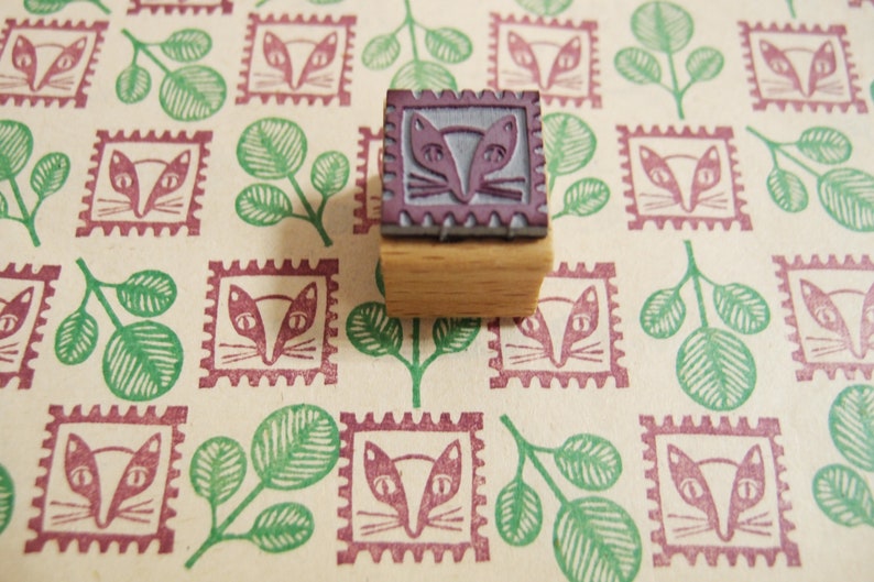 May include: A wooden stamp with a purple rubber design of a cat's face. The stamp is on a background of a repeating pattern of green leaves and burgundy cat faces.