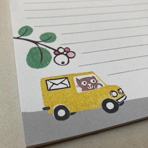 Könnte beinhalten: Notizblock mit linierten Seiten und einer skurrilen Illustration. Ein gelber Lieferwagen mit einem Katzenfahrer ist abgebildet, der Post transportiert. Ein grünes und rosa Blumenmuster befindet sich in der oberen linken Ecke. Der Notizblock liegt auf einer grauen Oberfläche.