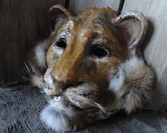 May include: A brown and white paper mache lion mask with fur accents around the eyes and mouth.
