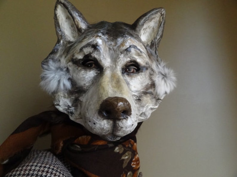 May include: A gray and white wolf mask with fur accents around the ears. The mask is made of paper mache and has realistic details, including a black nose and painted eyes.