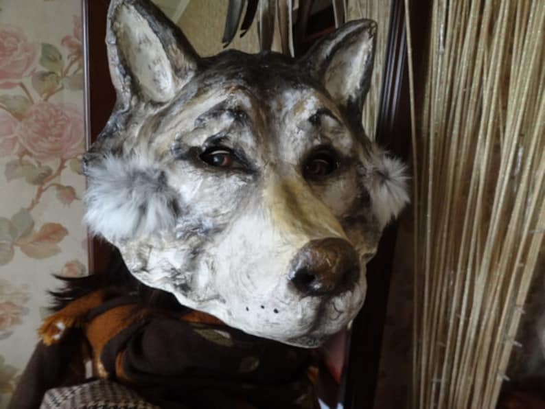 May include: A gray and white wolf mask with fur accents around the ears. The mask is made of paper mache and has a realistic look.