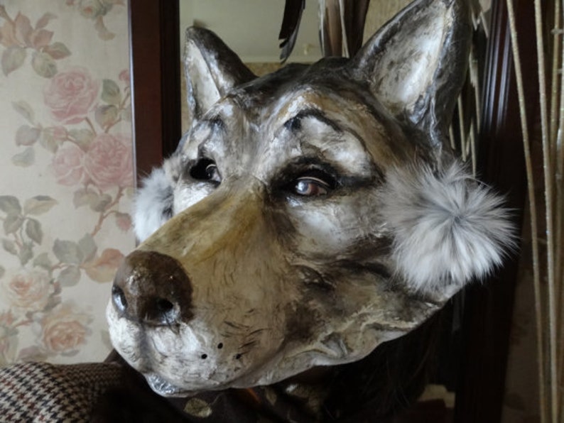 May include: A gray and brown wolf mask with white fur accents around the ears. The mask is made of paper mache and has realistic details, including a painted nose and eyes.