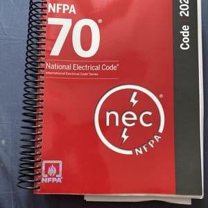 Peut inclure: Un livre à reliure spirale NFPA 70 National Electrical Code 2026 avec une couverture rouge. Le recto présente du texte blanc, un logo circulaire nec avec des éclairs et un petit emblème de flamme NFPA holographique. Une bande noire verticale est visible.