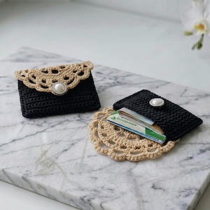 May include: Two rectangular crochet card holders in black yarn, each featuring a beige scalloped lace-style flap and a central white faux pearl button. One holder is closed, while the other is open and contains several plastic credit cards.