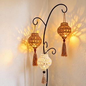 May include: Three hanging lanterns made of knotted cord, two in a tan shade and one in white, suspended from a black metal stand. Each lantern features a spherical shape, a wooden bead at the base, and a long tassel, casting warm light patterns.