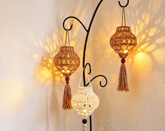 May include: Three hanging lanterns made of knotted cord, two in a tan shade and one in white, suspended from a black metal stand. Each lantern features a spherical shape, a wooden bead at the base, and a long tassel, casting warm light patterns.