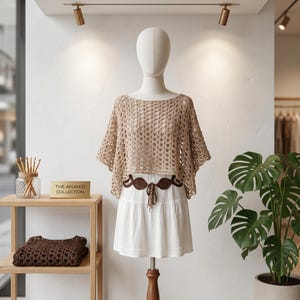 May include: A beige crochet poncho with a wide, open-weave pattern worn over a white skirt. A dark brown decorative belt with circular cutouts is tied at the waist. A sign on the shelf reads THE ANAHID COLLECTION.