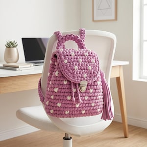 May include: A mauve crochet backpack featuring a repeating pattern of small white heart shapes. The bag has a top handle, a flap closure with a silver-toned metal clasp, and a long matching mauve tassel attached to the side of the main compartment.