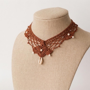 May include: A brown crochet choker necklace featuring an intricate openwork pattern, small white spherical beads integrated throughout the design, and a single off-white cowrie shell pendant hanging from the center of the lace-like structure.