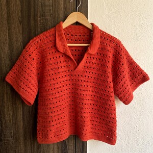 May include: A bright orange short-sleeved polo shirt crafted with an open-work crochet knit pattern. The garment features a classic collar, a V-neckline, and a straight hem, showcasing a textured, airy construction throughout the entire piece.