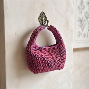 May include: A small, crescent-shaped handbag crocheted from variegated yarn in shades of magenta, pink, and deep purple. The bag features a thick, integrated handle and a textured, ribbed stitch pattern, hanging from a decorative metal wall hook.
