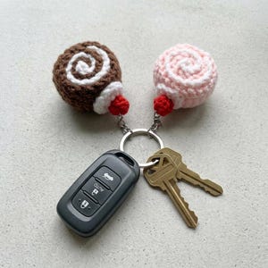 May include: A metal keyring holding a black car key fob, two brass keys, and two crocheted charms. One charm is a brown and white swirl, the other is a light pink and white swirl, each featuring a small red crocheted sphere at the base.