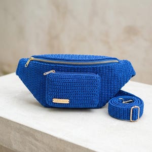 Puede incluir: Una riñonera de ganchillo en color azul real vibrante con un compartimento principal con cremallera y un bolsillo frontal más pequeño con cremallera. Incluye una correa de ganchillo ajustable a juego con herrajes metálicos dorados y una etiqueta dorada SULIS.