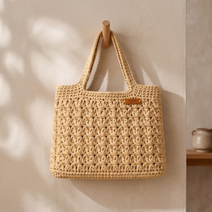 Puede incluir: Un bolso tote de ganchillo en color beige con un patrón repetitivo de puntos de concha y dos asas resistentes. Una pequeña etiqueta rectangular de cuero marrón está unida al lado superior derecho del bolso, mostrando la palabra Handmade en relieve.