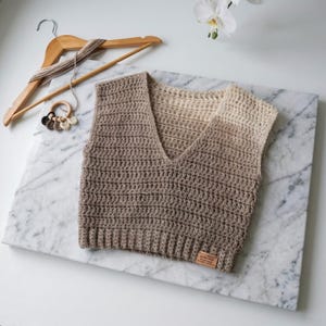 May include: A sleeveless crochet vest featuring a V-neckline and a gradient colour pattern that transitions from dark brown at the bottom to cream at the shoulders. The garment includes a small rectangular leather tag at the bottom hem.