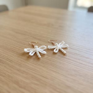 Puede incluir: Un par de pendientes con forma de libélula hechos con hilo blanco y pequeñas cuentas blancas. Cada pendiente presenta un delicado diseño de alas de ganchillo y un cuerpo de cuentas, suspendido de un sencillo gancho de metal dorado.