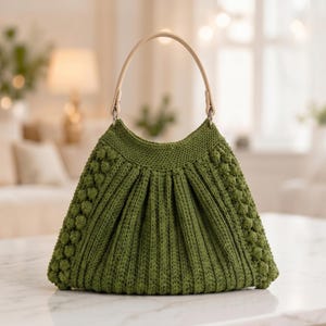May include: A sage green crocheted handbag featuring a textured bobble stitch pattern along the sides and vertical ribbed detailing across the front. The bag includes a single, smooth, light beige arched handle attached with silver metal rings.