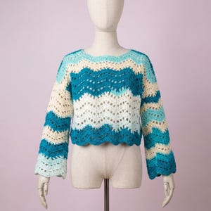 Puede incluir: Un suéter de ganchillo de manga larga con un patrón de chevrón horizontal en tonos alternos de azul cerceta, azul claro y crema. La prenda tiene un cuello redondo, un corte corto y un dobladillo festoneado con una textura de punto abierto.