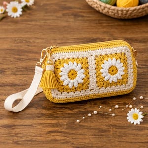 May include: A rectangular crochet pouch featuring two square motifs with white daisy patterns on a yellow background. The pouch has a cream-coloured wrist strap, a gold-tone metal clasp, and a matching yellow yarn tassel attached to the side.