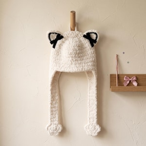 May include: A cream-coloured crocheted hat featuring cat-like ears with black inner accents and small black bows. The hat includes two long side flaps ending in paw-shaped details. A pink crochet hook and a small pink bow are visible nearby.