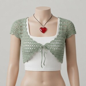 May include: A sage green crochet bolero with short sleeves and a front tie closure, worn over a white ribbed tank top. A red heart-shaped pendant hangs from a thin black cord around the neck, completing the layered look of this garment.