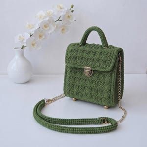 May include: A structured olive green crochet handbag featuring a textured stitch pattern, a top handle, and a metallic push-lock closure. The bag includes a detachable gold-tone chain strap with a matching crochet shoulder pad and four small metal feet.