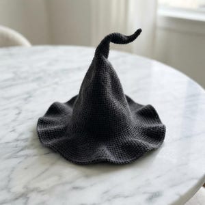May include: A dark charcoal gray crochet hat featuring a tall, pointed, and slightly curved crown with a wide, wavy brim. The hat is constructed from a dense, uniform stitch pattern, creating a textured surface throughout the entire piece.