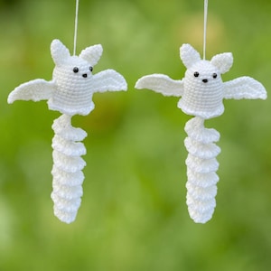 May include: Two white crocheted bat ornaments hanging from thin strings. Each bat features a rounded head with pointed ears, small black bead eyes, outstretched wings, and a long, spiraling tail extending downward from the body.