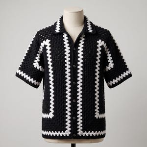 May include: A short-sleeve button-down shirt crafted from black and white crocheted yarn. The garment features a prominent white crocheted border along the collar, the front button placket, the sleeve cuffs, and the hem, creating a geometric pattern.