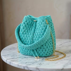 May include: A bright turquoise crochet bucket bag featuring a textured, woven pattern. The bag includes a matching crochet handle and a long, thin gold-tone metal chain strap. A drawstring closure is integrated into the top of the bag.