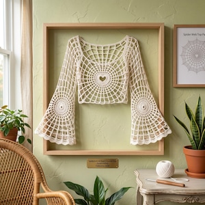 Puede incluir: Un top de ganchillo color crema con un patrón de telaraña, un recorte central en forma de corazón y mangas largas acampanadas. La prenda se exhibe dentro de un marco de madera, con una placa que dice Spiderweb & Heart Crochet Top - Hand-crafted - Collection 2024.