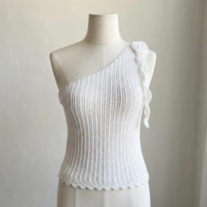 May include: A white one-shoulder crochet top featuring a vertical ribbed stitch pattern, a decorative floral accent on the shoulder strap, and a scalloped edge along the bottom hem, displayed on a neutral-colored mannequin torso.