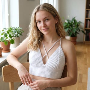Op de afbeelding: Een wit gehaakte crop top met een diepe V-hals en gekruiste bandjes die over de schouders lopen. Het kledingstuk is gemaakt met een gestructureerd ribbelpatroon over het hele lijfje, wat zorgt voor een nauwsluitende pasvorm.